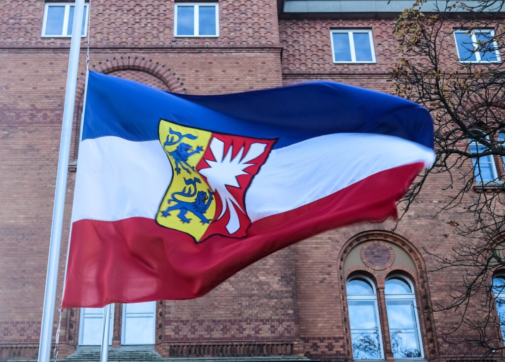 Flagge mit blau-weiß-roten Streifen und Wappen, weht vor Ziegelgebäude - Symbol für Solidarität und faire Bestattungen.