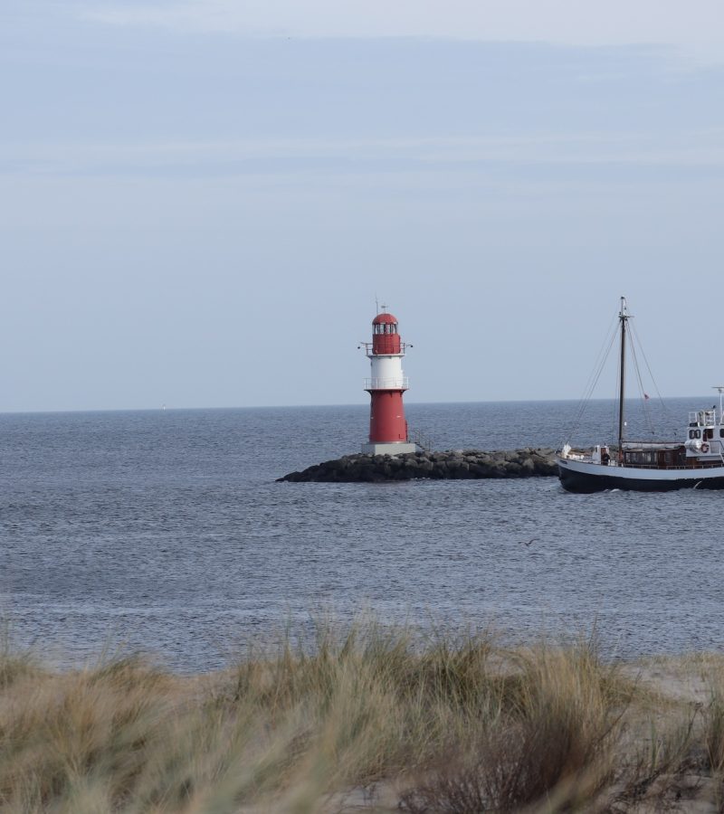 Roter und weißer Leuchtturm am Meer mit Fischerboot - Symbol für Orientierung, Sicherheit und Abschied bei YANA Bestattung.