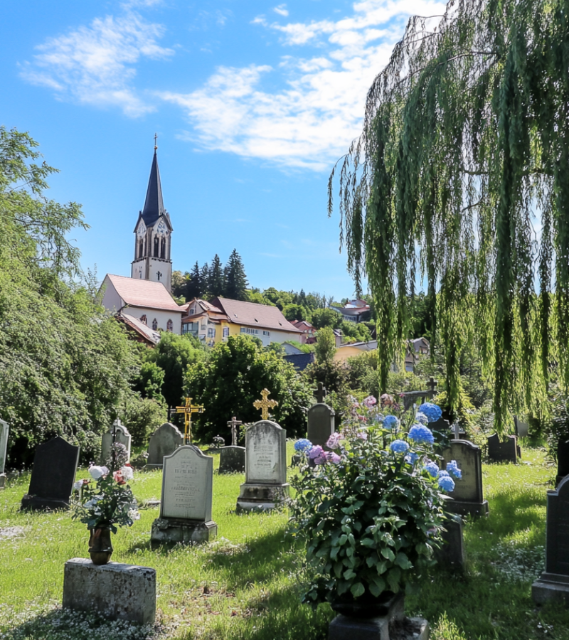 Grüner Friedhof mit Grabsteinen und Blumen, im Hintergrund Kirche - würdevolle Bestattung in Bayern.